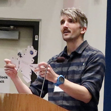 Kyle Ormsby at a lectern holding a paper model of an associahedron